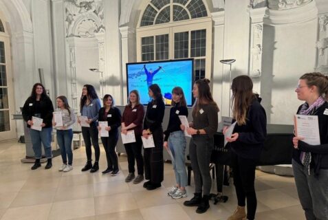 10 Frauen stehen in einem festlichen Raum im Halbkreis vor einem Flügel. Alle Frauen halten ein Blatt Papier in der Hand.